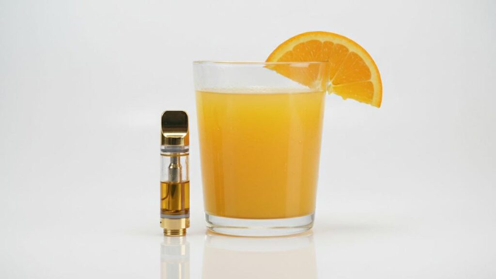 A golden cannabis vape cartridge, fresh orange juice, and an orange slice on a clean white background.